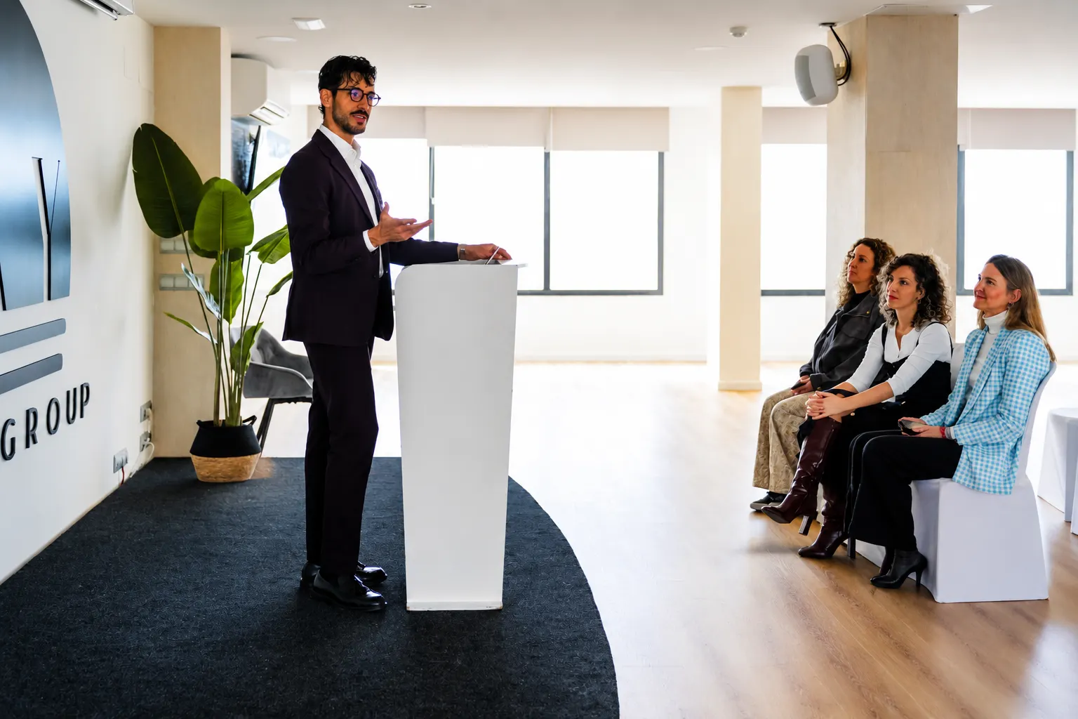 En una sala de conferencias moderna y luminosa de Fay Victoria Beach, un hombre con gafas y un traje oscuro se encuentra de pie frente a un podio blanco, gesticulando mientras habla con confianza.